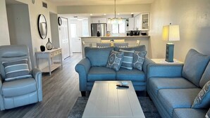 A modern living room with a grey sofa, a white coffee table, and a grey armchair. There is a kitchen in the background with white cabinets and a chandelier. - Runaway Bay 107 - Beach, Pool, gym and much more (Unknown)