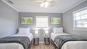 Een slaapkamer met twee bedden, een plafondventilator, een raam met zonwering en twee aan de muur hangende schilderijen van palmbomen.