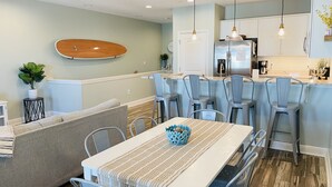 A modern kitchen with a dining area, featuring a wooden surfboard on the wall, a striped tablecloth, and a blue crocheted bowl. - Family Friendly, Close to Beach and Shops (Destin)