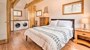 Un dormitorio con cama, cabecera de madera, mesita de noche, cómoda, ventana y zona de lavandería con lavadoras.