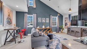 Ein modernes Wohnzimmer mit grauem Sofa, einem schwarz-roten Barhocker und einem Holztisch. Ein großes Fenster mit Vorhängen, eine Küchenzeile mit Marmorarbeitsplatte und Kunstwerken an den Wänden.
