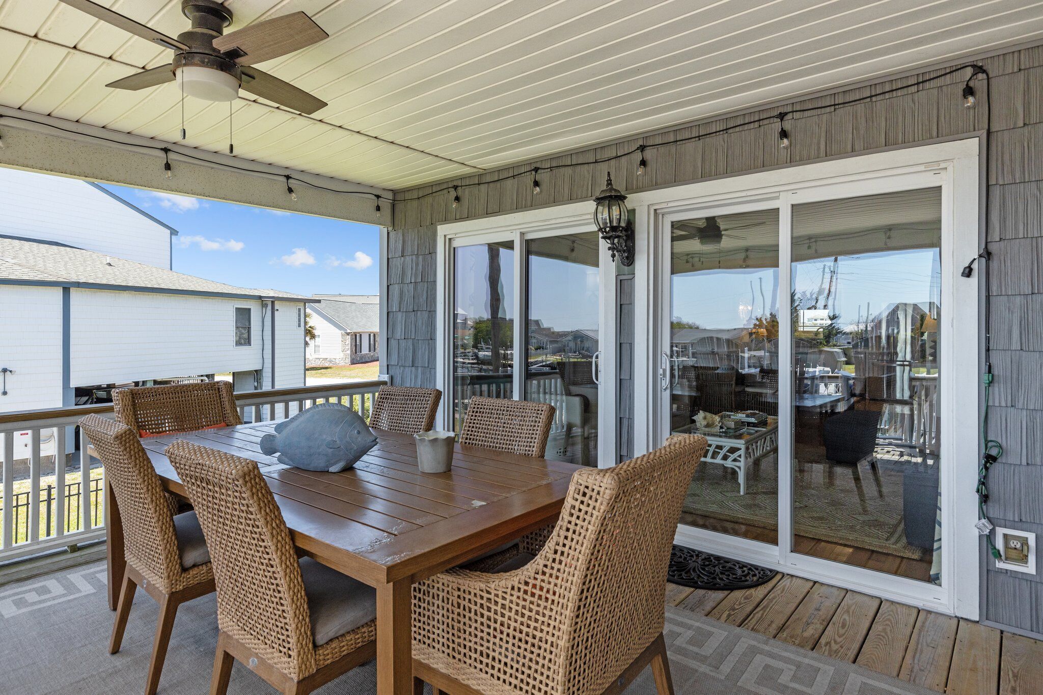 Eine überdachte Terrasse mit Holztisch und Korbstühlen, großen Schiebeglastüren und einem Deckenventilator.
