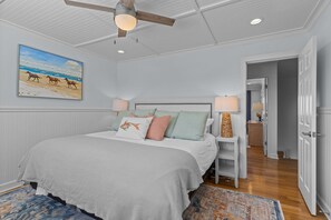 Une chambre à coucher avec un lit, un ventilateur de plafond, un tableau représentant des chevaux accroché au mur et une porte donnant sur une autre pièce.