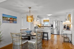 A modern kitchen with white cabinetry, a stainless steel refrigerator, and a dining area with wicker chairs and a table. - Waterfront Paradise! Boatlift+ Sunset views! (Atlantic Beach)