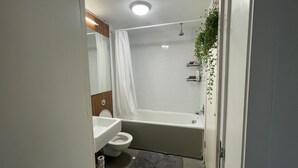 Ein modernes Badezimmer mit einem weißen Waschbecken, einer Toilette und einer Badewanne. Ein Duschvorhang, eine kleine Pflanze und ein grauer Teppich liegen auf dem Boden.