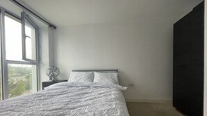Ein Schlafzimmer mit einem großen Bett, einem Nachttisch, einem Ventilator und einem Fenster mit Blick auf Gebäude und Bäume.
