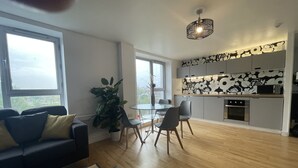 Ein modernes Wohnzimmer mit einem Glastisch, einem Schachspiel und einer Couch mit Kissen. Es gibt einen Essbereich mit einem Glastisch und Stühlen sowie eine Küche mit Mikrowelle und Backofen.