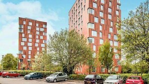 Ein modernes, mehrstöckiges Gebäude mit Ziegelaußenwand und zahlreichen Fenstern. Vor dem Gebäude sind mehrere Autos geparkt.