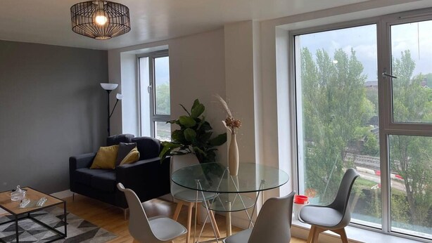 Ein modernes Wohnzimmer mit einem Glastisch, grauen Stühlen, einem schwarzen Sofa und einem großen Fenster mit Blick auf Bäume und Gebäude.