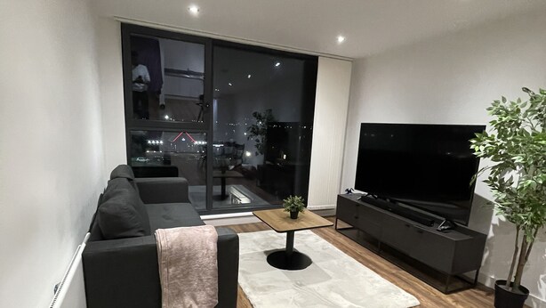 A modern living room with a black sofa, a coffee table, a TV, and a large window overlooking a cityscape. - Luxury 2-bed Apartment in Manchester With Parking (Salford)