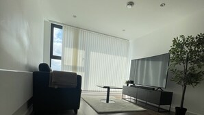 A modern living room with hardwood flooring, a dark armchair, a flat-screen TV, a potted plant, and a large window with blinds. - Luxury 2-bed Apartment in Manchester With Parking (Salford)