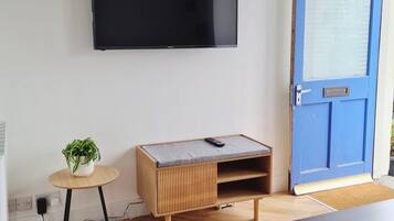 A room with a flat-screen TV mounted on the wall, a blue door, a wooden cabinet with a mattress, a small round table with a plant, and a dark wooden table.