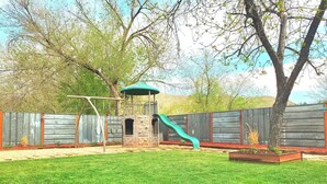 A backyard with a play structure, a green slide, a fenced area, and a tree. - Comfortable, Laid-back Cottage. Private Yard. Near Zion. 2 Bedroom, Full Kitchen (Hurricane)