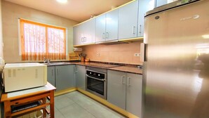 Une cuisine compacte avec des meubles gris, un réfrigérateur en acier inoxydable, un four encastré et un micro-ondes posé sur une table en bois.