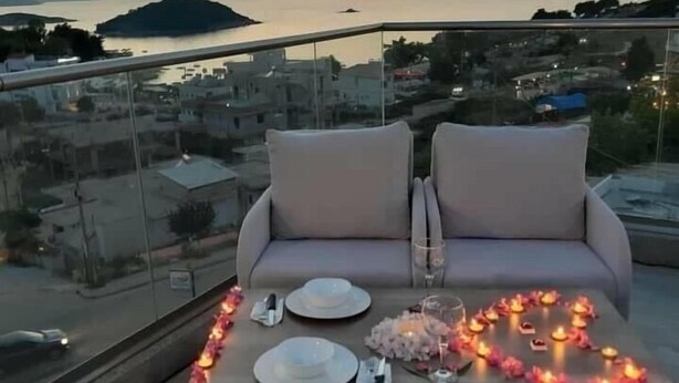A balcony with a table set for two, overlooking a cityscape and water at sunset. - La Luna Resort Ksamil (Ksamil)