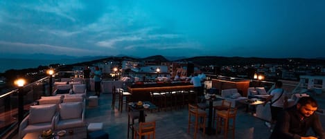 Un bar en la azotea con mesas al aire libre, una zona de bar y vistas a la ciudad al anochecer.
