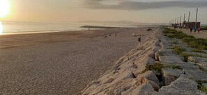 Ein Strand mit einer Felsbucht, einige Spaziergänger und ein Sonnenuntergang im Hintergrund.
