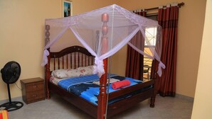 Een houten hemelbed met een klamboe, een ventilator naast het bed en een bloemenprint sprei.