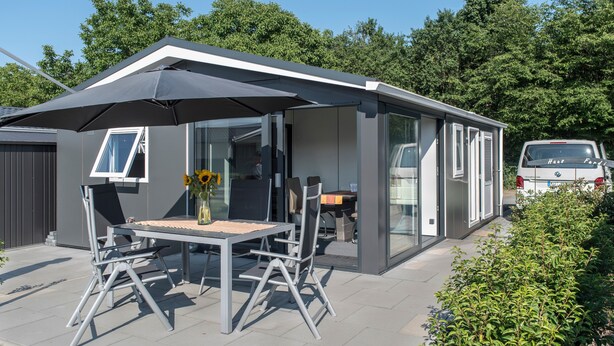 Une terrasse extérieure moderne, dotée d’une table et de chaises, d’un grand parasol et d’une pièce fermée par des baies vitrées offrant une vue sur les environs.
