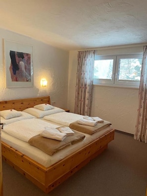 A hotel room with a wooden bed, a painting on the wall, a window with curtains, and a wall-mounted light. - Apartment 'W8 Haus Ganghofer' with Mountain View, Balcony & Wi-Fi (Leutasch)