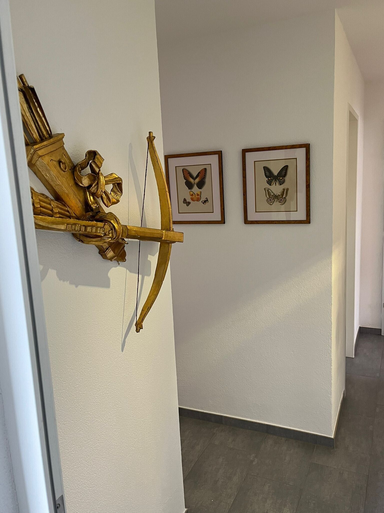 Un pasillo con un arco y una flecha dorados en la pared, dos cuadros con imágenes de mariposas y un piso de baldosas.