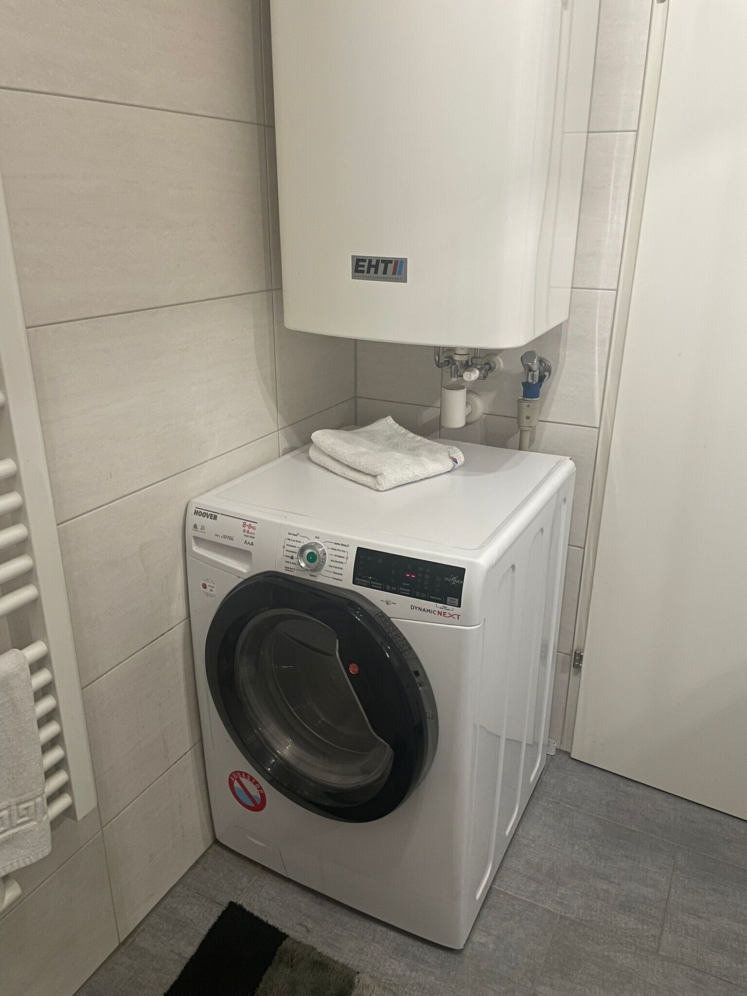 Una lavadora blanca compacta de carga frontal, con panel de control, ubicada en un baño pequeño con azulejos, un inodoro blanco y un perchero.