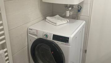 Una lavadora blanca compacta de carga frontal, con panel de control, ubicada en un baño pequeño con azulejos, un inodoro blanco y un perchero.