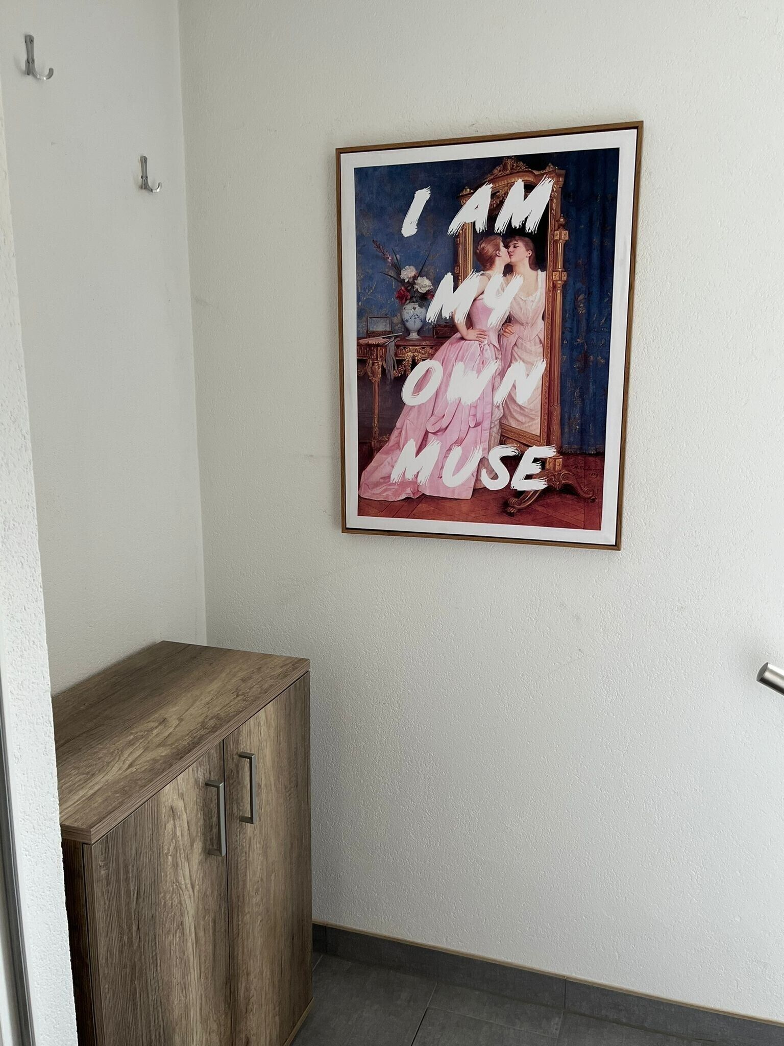 Une œuvre d’art encadrée, sur laquelle on peut lire le texte « JE SUIS MA PROPRE MUSE », est accrochée à un mur blanc.