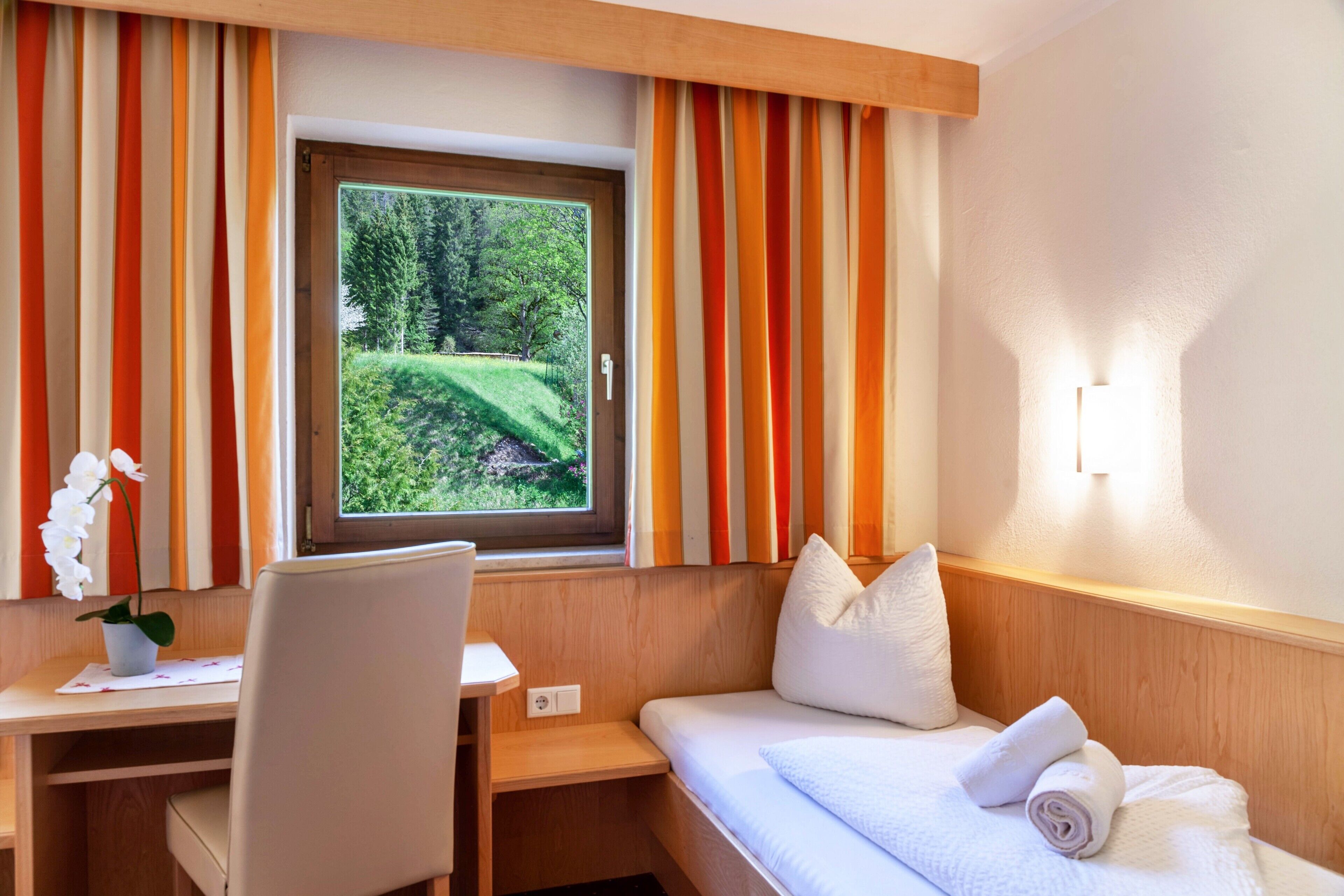 A hotel room with a bed, a desk, a chair, a window with curtains, and a view of greenery outside.