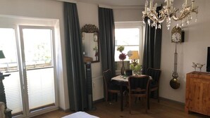 Une pièce comprenant un lit, une table à manger avec des chaises, un lustre, une horloge et une fenêtre avec des rideaux.