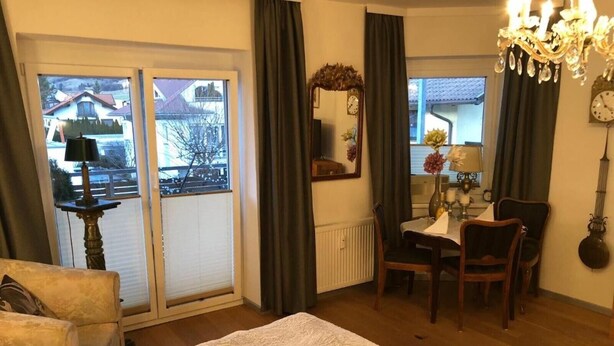 Une chambre d’hôtel avec un lit, un bureau et une chaise, une fenêtre avec des rideaux, un lustre et une horloge au mur.