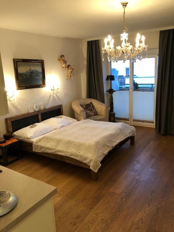 Une chambre d’hôtel avec un lit, une chaise, un bureau, un lustre, un tableau et une fenêtre avec des rideaux.