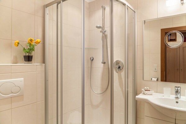 Un baño moderno con ducha acristalada, lavamanos con espejo y una planta en maceta con flores amarillas.