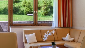 A room with a sofa, a window with curtains, and a view of greenery outside. - Apartment W3 Haus Ganghofer with Mountian View, Shared Terrace & Wi-Fi (Leutasch)