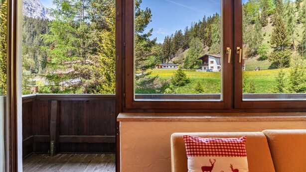 A room with a wooden deck, a sofa with a checkered pillow, and large windows offering a view of a mountainous landscape. - Apartment W4 Haus Ganghofer with Mountain View, Balcony & Wi-Fi (Leutasch)