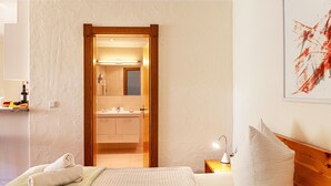 Hotelkamer met een bed, nachtkastje met een lamp, houten hoofdeinde, wanddecoratie en een badkamer zichtbaar door een geopende deur.