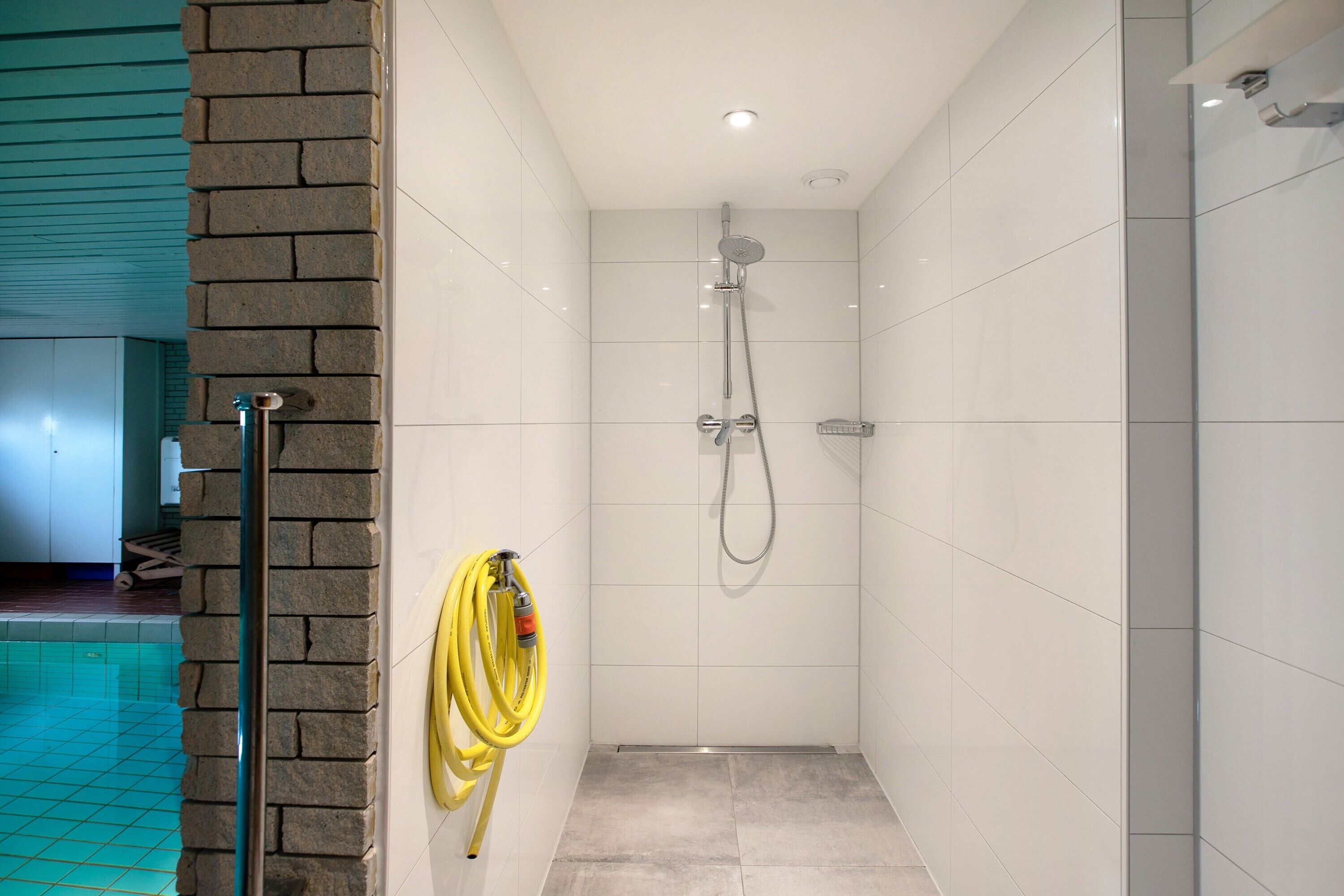 Ducha con manguera, pared de azulejos y piscina al fondo.