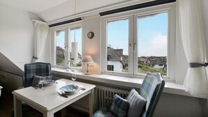 A dining area with a table set for two, a window with curtains, a lamp, and a view of houses outside. - Holiday Apartment 'Haus Lazy Dolphin No. 10' with Sea View and Wi-Fi (Rantum)