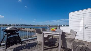 Eine Terrasse mit Grill, Korbstühlen, einem Tisch mit Obst und Blick auf das Wasser.