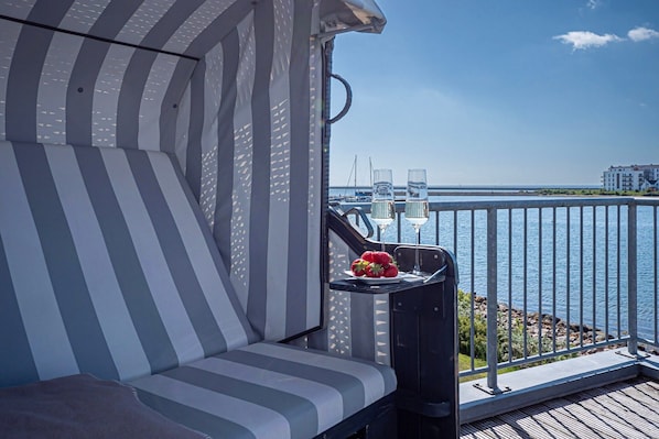 A balcony with a striped sofa, a small table with a glass of water and a plate of strawberries, and a view of the sea and buildings.