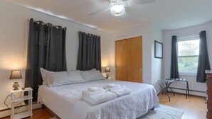 Ein ordentlich bezogenes Bett mit weißen Leinen und Handtüchern, ein Deckenventilator, ein kleiner Nachttisch mit Lampe und ein Fenster mit Vorhängen.