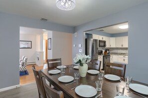 Un coin repas avec une table en bois dressée pour quatre personnes, un vase rempli de fleurs et une vue sur la cuisine, avec ses armoires blanches et son réfrigérateur.