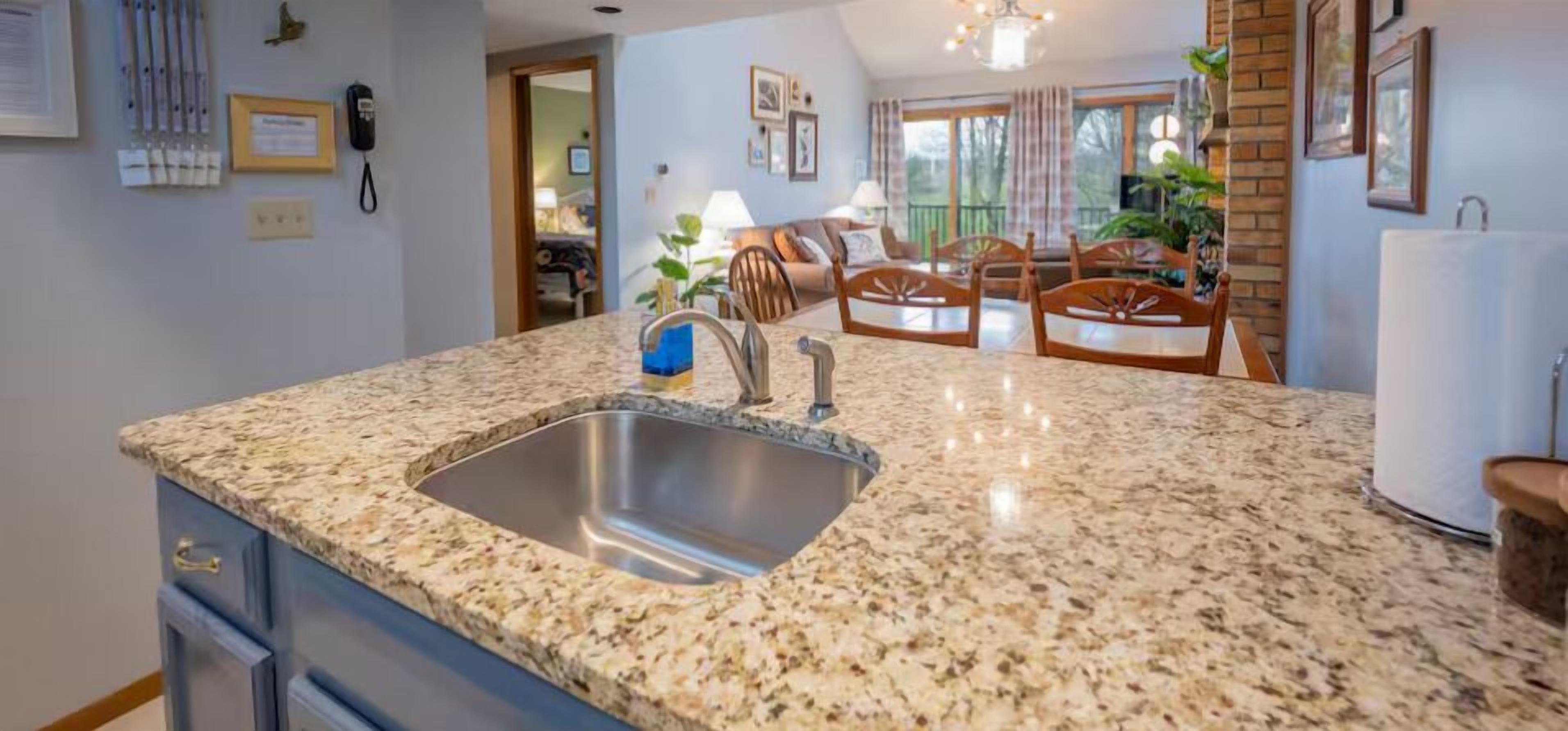 A kitchen with granite countertops, a stainless steel sink, and wooden chairs.