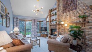 Ein Wohnzimmer mit Kamin aus Backstein, zwei Sofas, einem Fernseher und einem Balkon mit Vorhängen.