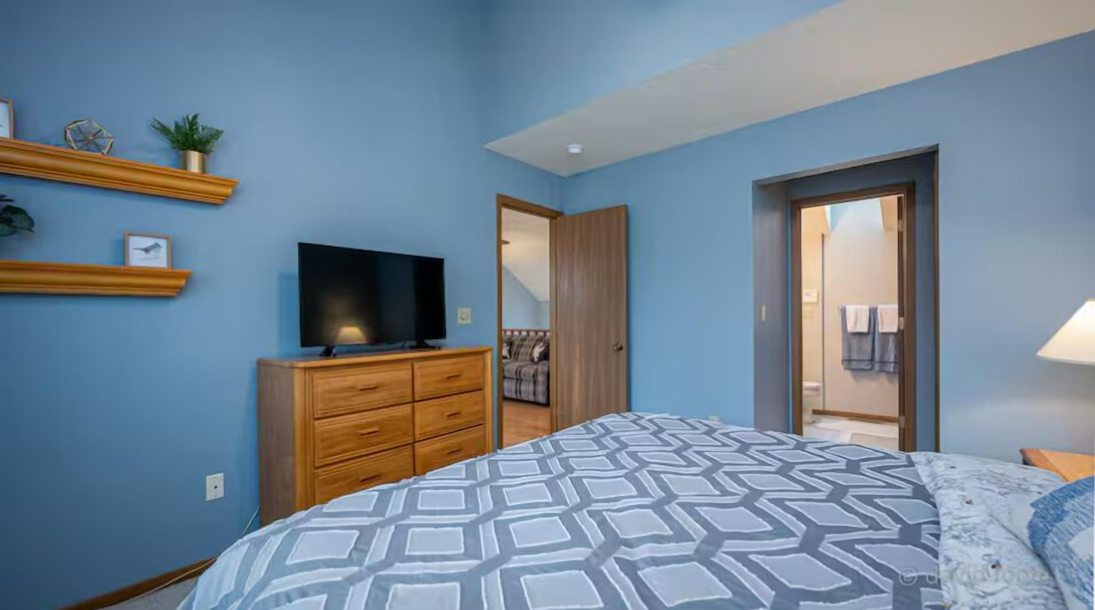 A bedroom with a bed, a TV on a wooden dresser, and a bathroom visible through an open door.