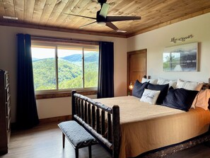 A bedroom with a wooden headboard, a bed with multiple pillows, a bench, a ceiling fan, a window with curtains, and a wall-mounted picture.