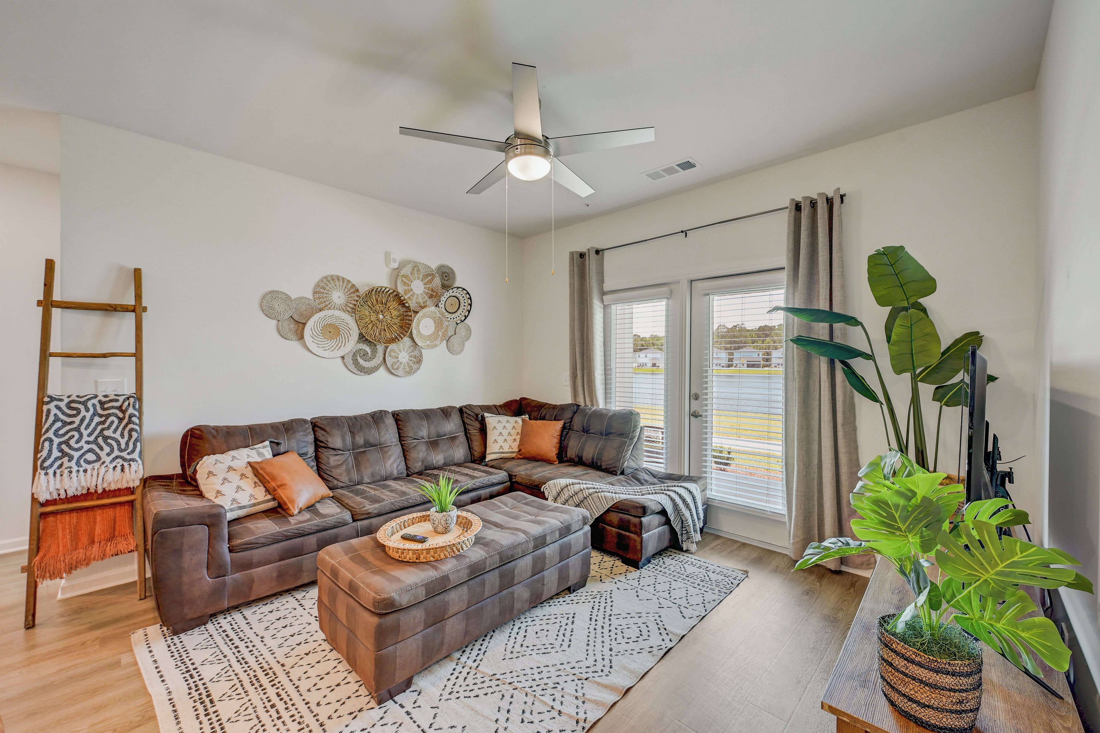 A modern living room with a sectional sofa, a ceiling fan, a decorative wall piece, and a large plant.