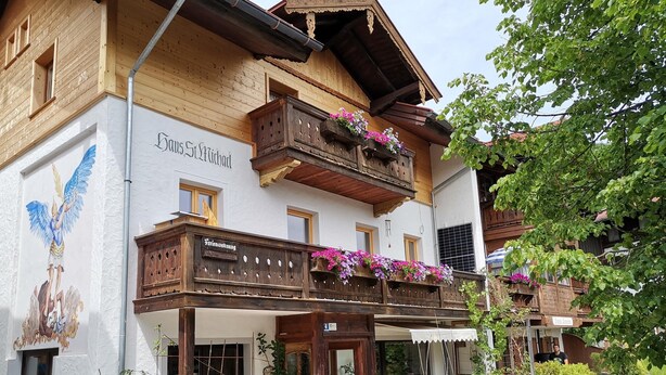 Ein traditionelles Holzhaus mit Balkonen, die mit Blumen geschmückt sind, einem Schild mit der Aufschrift "Gasthof St. Michael" und einem Wandgemälde eines Engels.