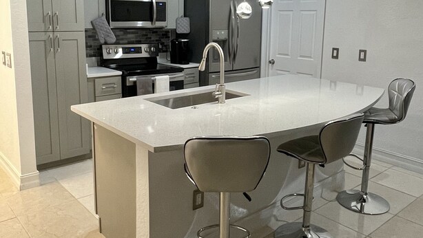 A modern kitchen with grey cabinets, stainless steel appliances, a large island with bar stools, and pendant lighting.