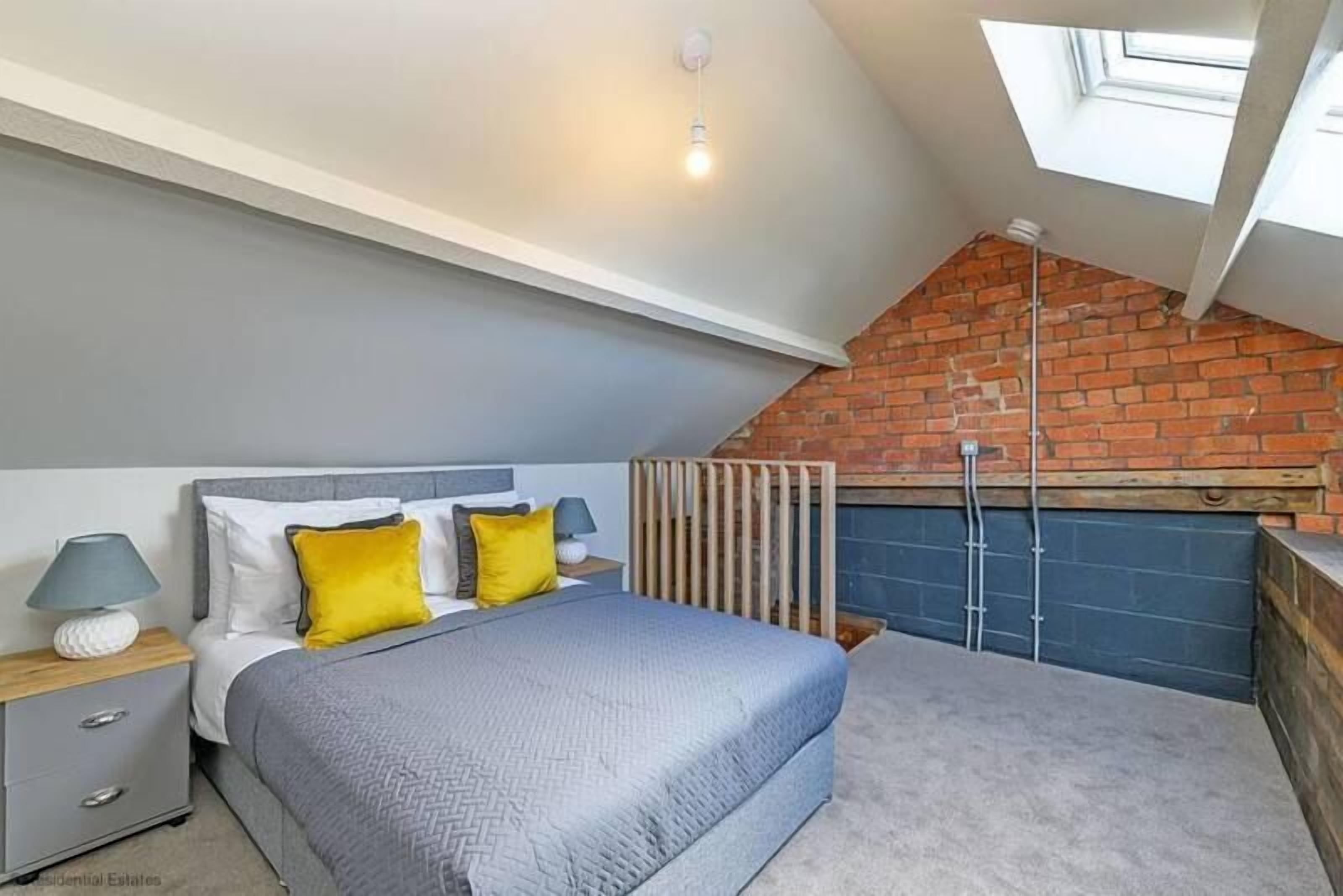 A bedroom with a sloped ceiling, a bed with a grey quilt, two yellow pillows, a bedside table with a lamp, and a view of a brick wall and a window.